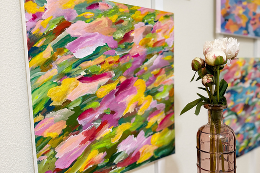 Vibrant abstract paintings full of shapes and colors hanging on a wall and a vase of flowers sitting in front.  